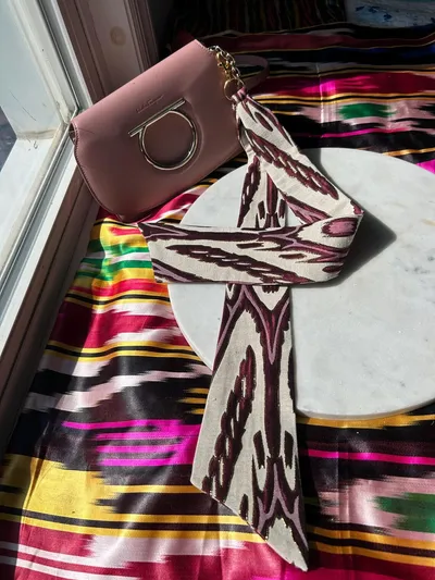 Pink bag with ikat ribbon charm styled against a window, with colorful ikat fabric draped alongside