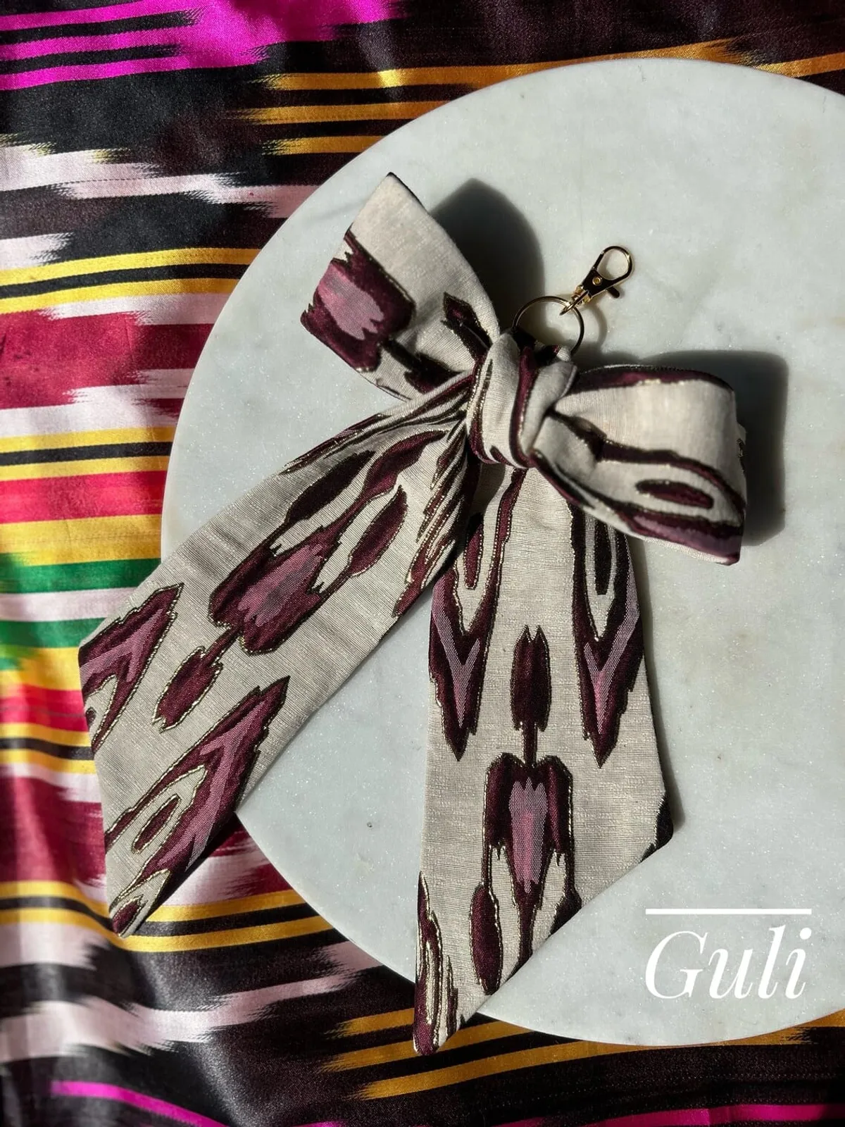 Atlas ikat silk ribbon bow bag charm with gold lobster clasp, in cream and burgundy, laid on a marble round against a striped ikat backdrop