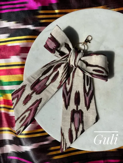 Atlas ikat silk ribbon bow bag charm with gold lobster clasp, in cream and burgundy, laid on a marble round against a striped ikat backdrop