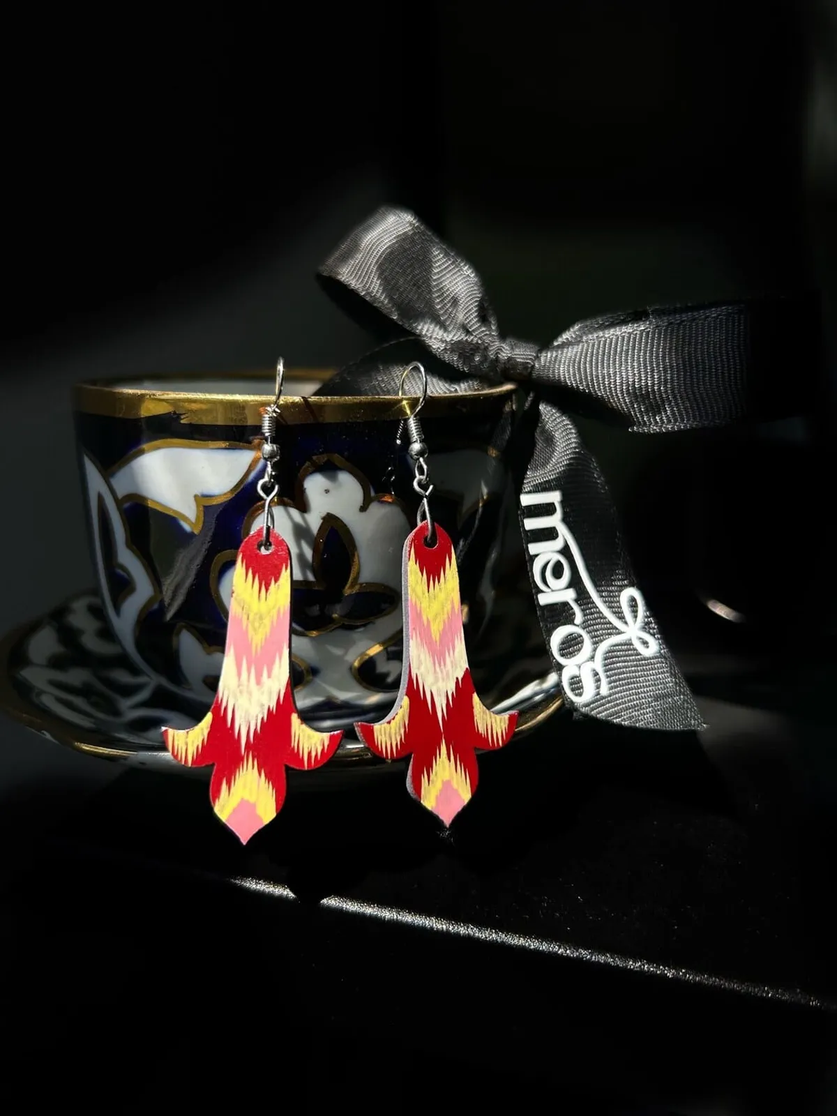 Red, yellow, and pink ikat-printed earrings hanging from a vintage teacup with a Meros ribbon