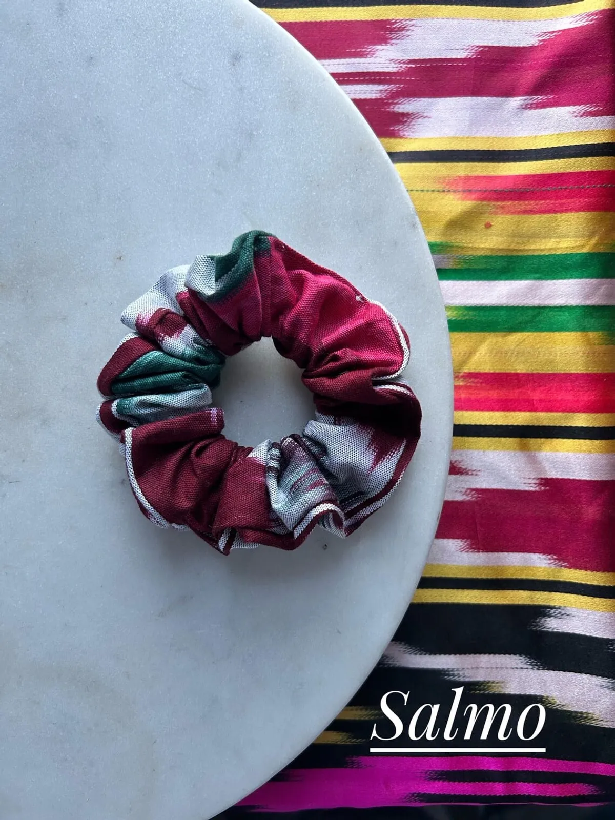 A maroon, white, and green plaid ikat scrunchie on a marble plate, labeled 'Salmo'