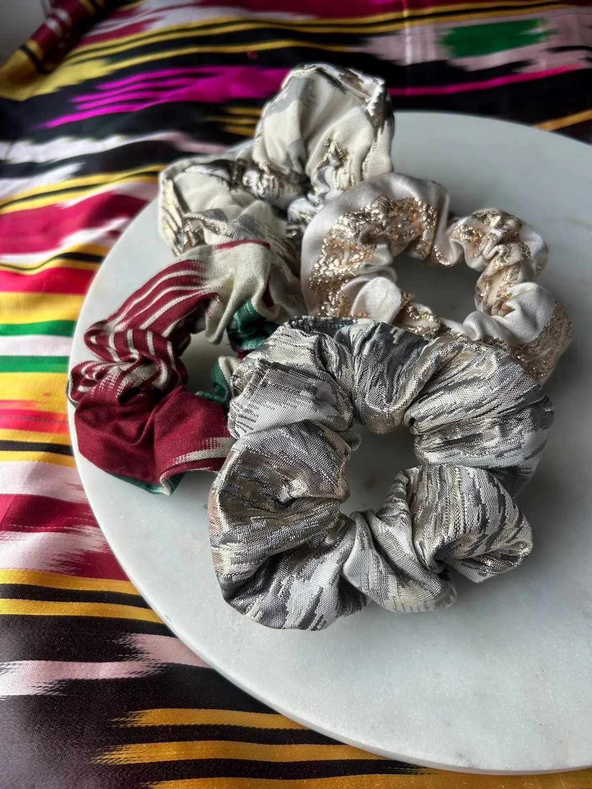 A pile of atlas ikat scrunchies in silver, red-and-white, and gold-embellished colorways on a marble plate