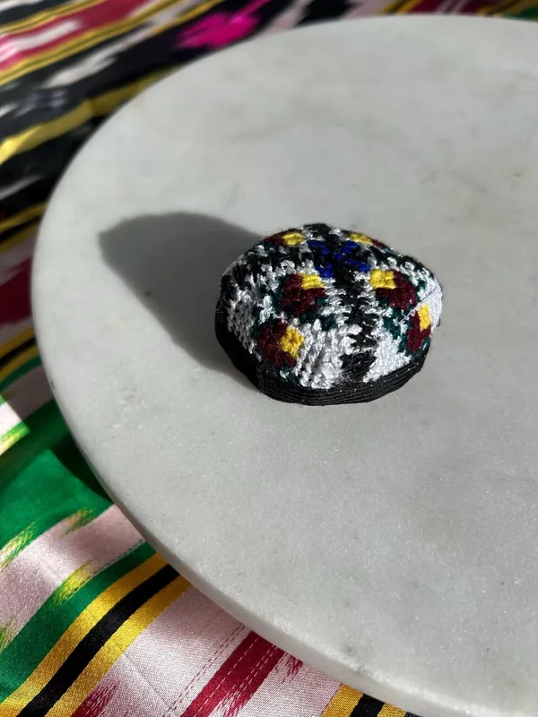 A small embroidered doppi-style hair tie in a black, white, red, and yellow cross-stitch pattern on a marble plate