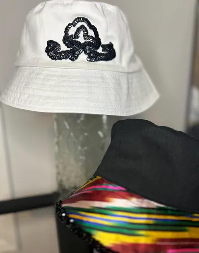 A white bucket hat with a black sequin swirl emblem, with a black ikat-trimmed bucket hat visible behind it