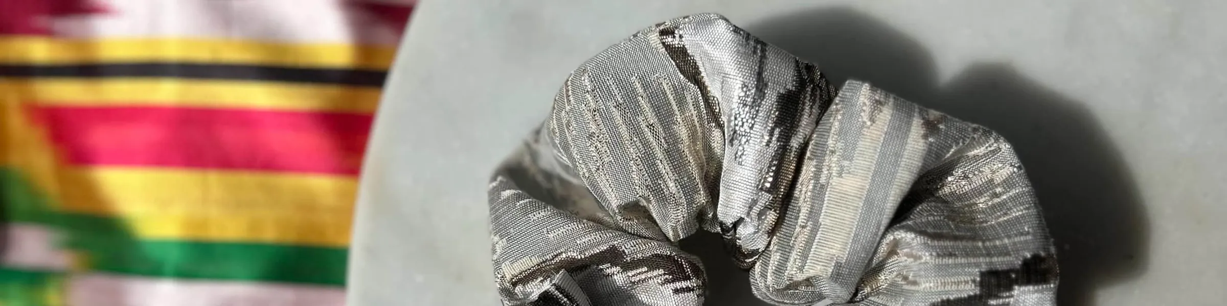 A silver atlas ikat scrunchie on a marble plate, with striped ikat silk fabric in the background